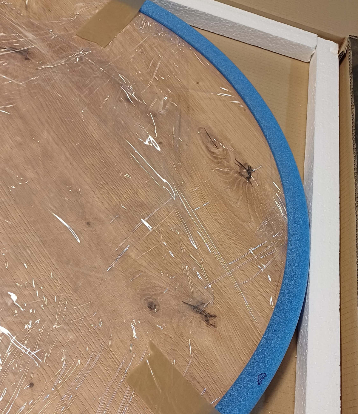 Round oak tabletop wrapped in film, its edge protected with foam and polystyrene, in a carton